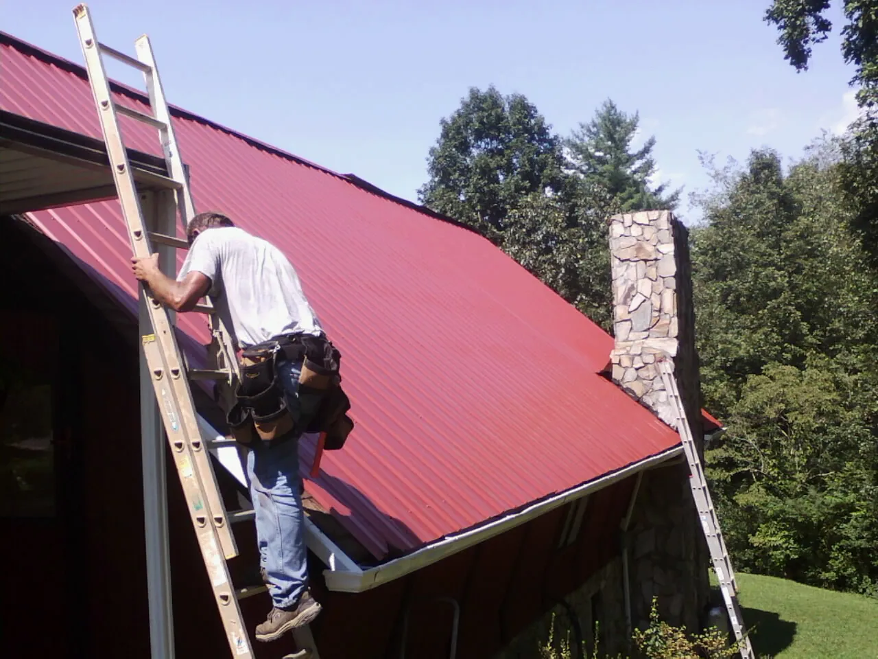 Roofer on a ladder beside a red metal roof and stone chimney — Soffit & Fascia Repair by Roofing Brothers in Fossil